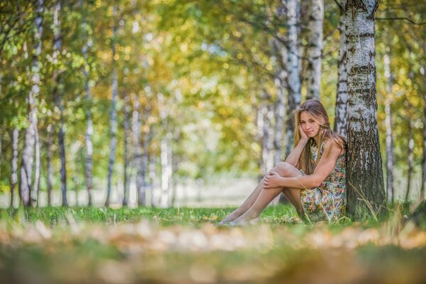 Chica en vestido sentado junto a un árbol en el bosque