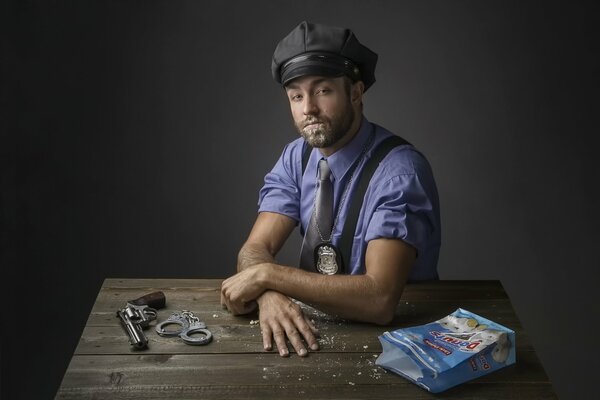 Un policía posando para una foto. Sobre la mesa hay un revólver, esposas y una bolsa de donas